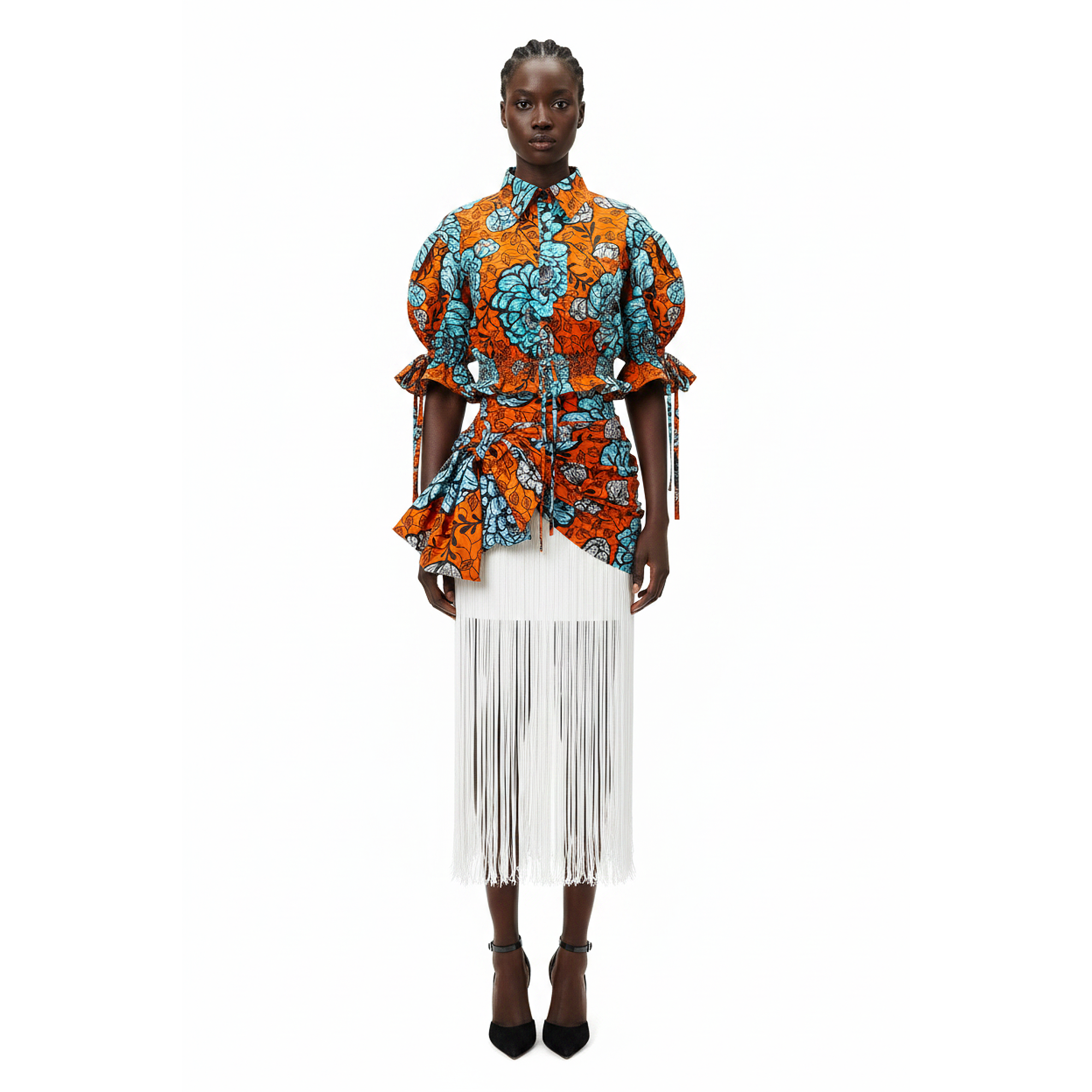 A model stands against a solid white background wearing a vibrant orange and teal patterned short-sleeve blouse with ruffled details, paired with a white fringed midi skirt and black high heels.