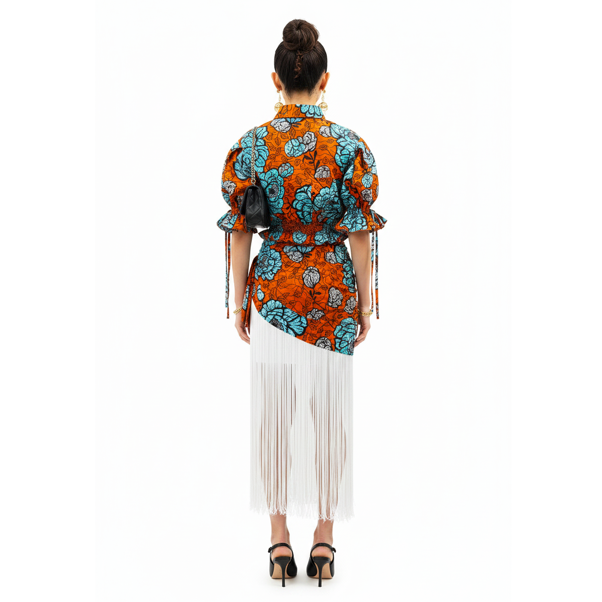 A woman viewed from the back wearing a bright orange and turquoise patterned wrap-style top with puff sleeves and a white fringe skirt, paired with black heels, against a plain white background.
