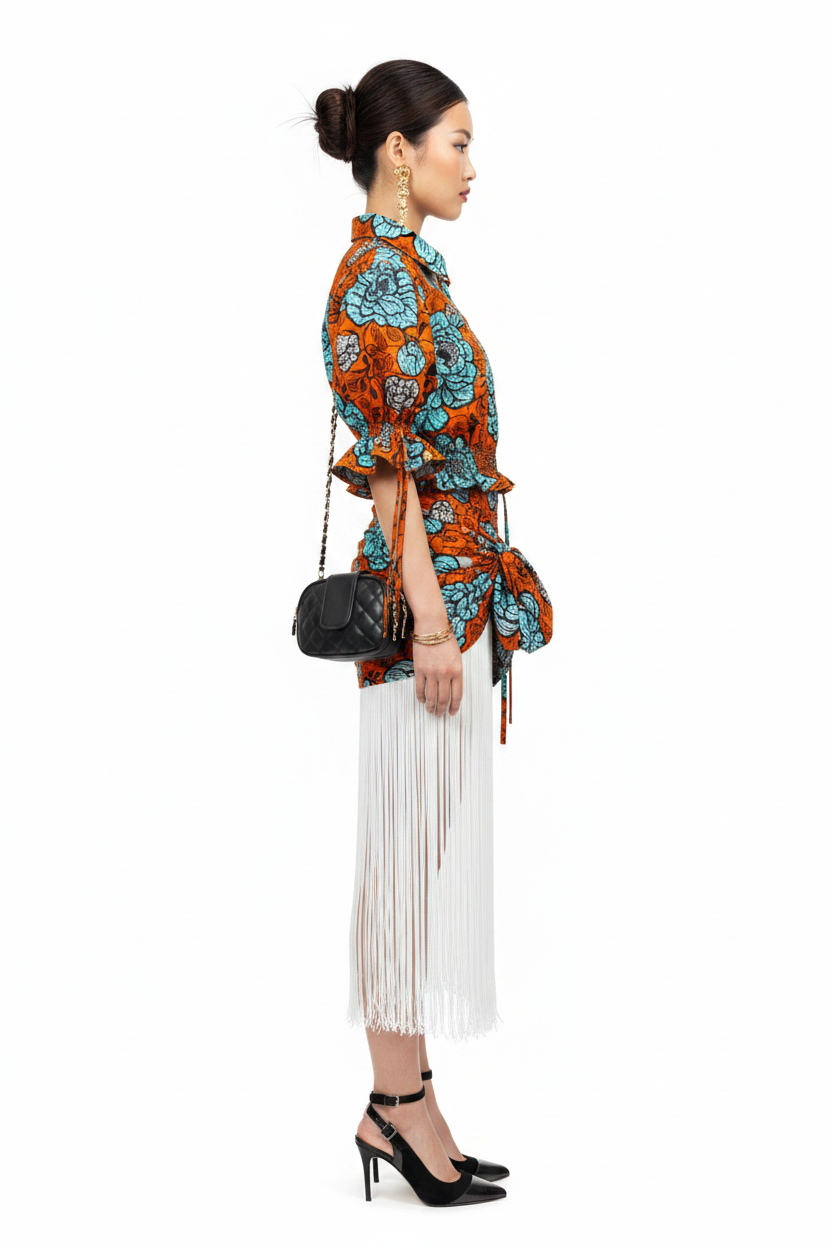 A woman in profile wearing a vibrant orange and turquoise patterned shirt with a fringed white skirt, black ankle-strap heels, and a small black quilted shoulder bag, set against a plain white studio background.