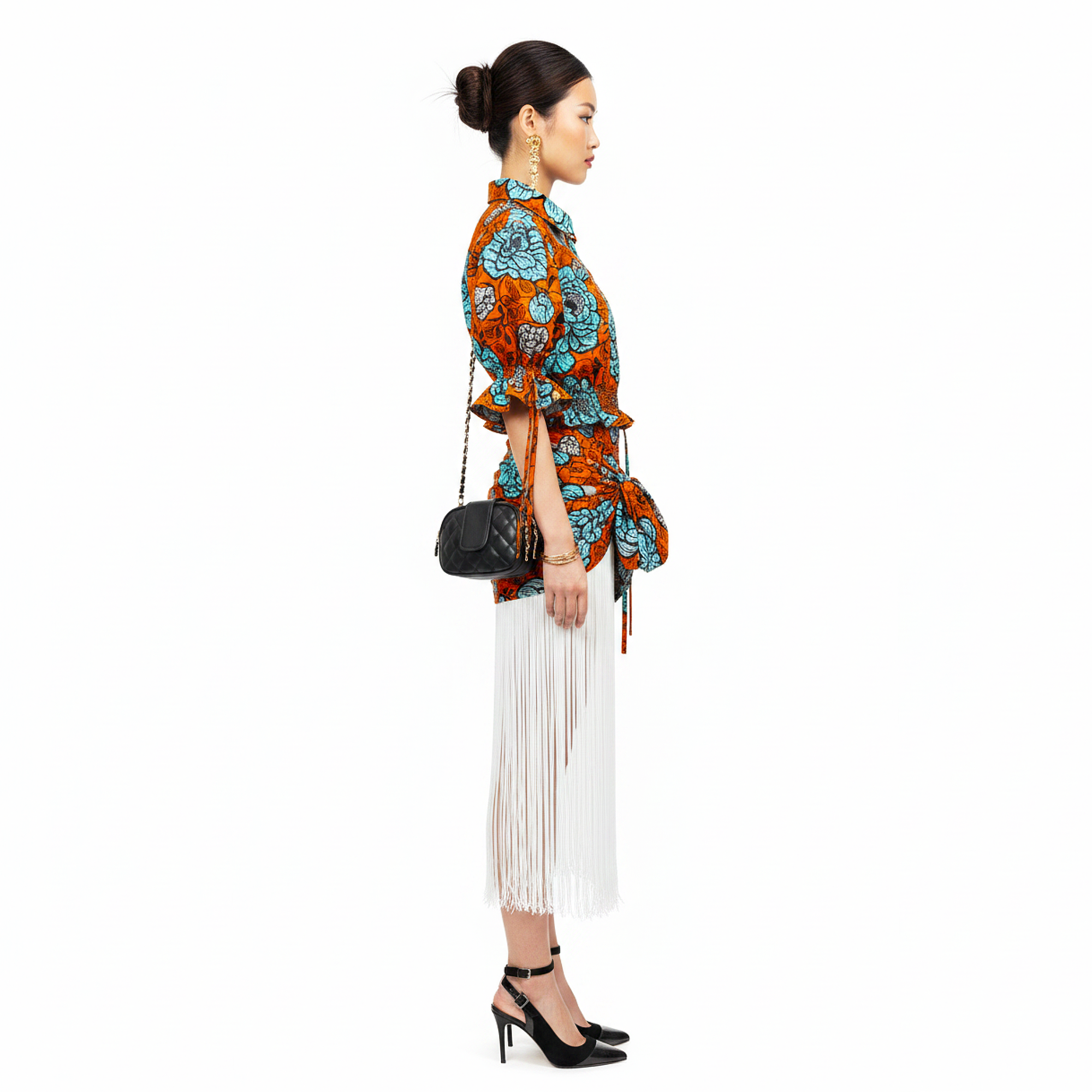 A model is shown in profile wearing a vibrant orange, blue, and black patterned shirt with a matching belt, paired with a long white fringe skirt and black high heels, carrying a small black shoulder bag against a solid white background.