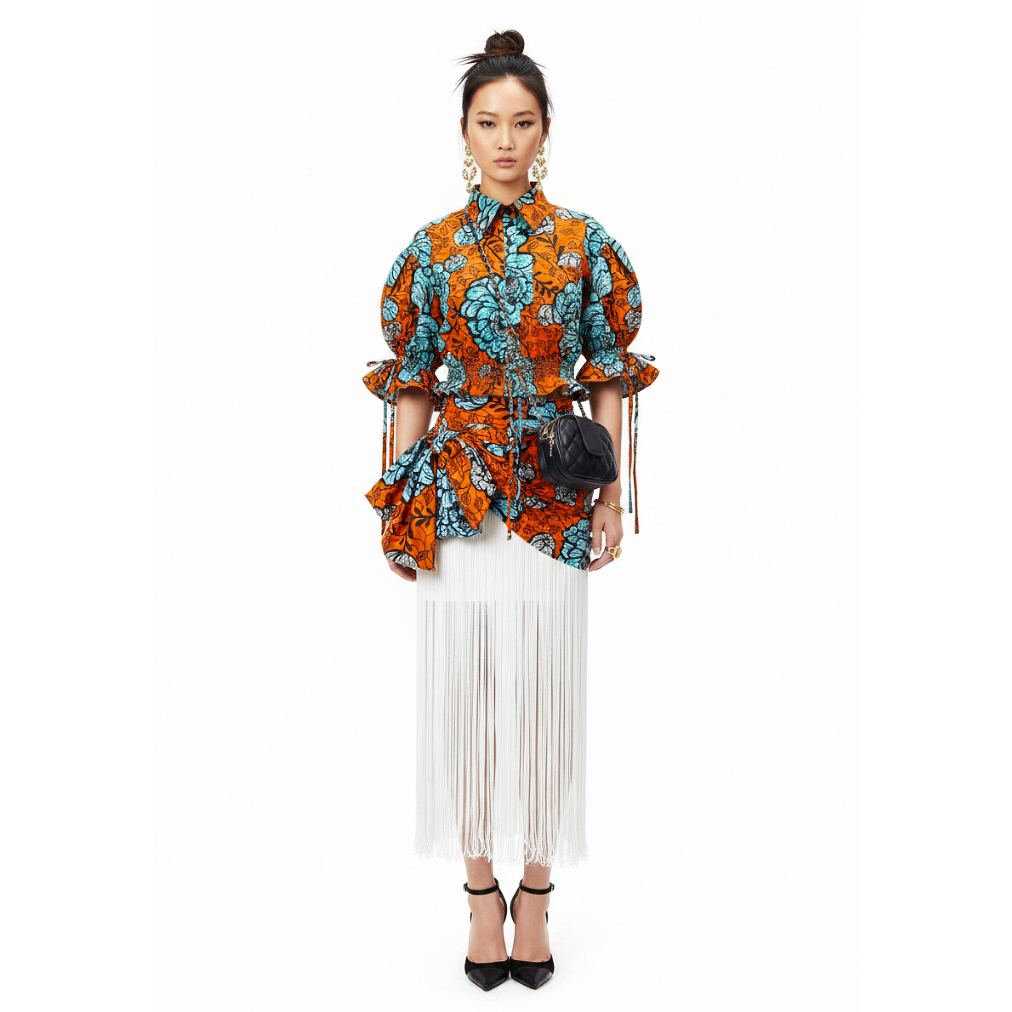 A model stands against a solid white background wearing a vibrant orange and turquoise patterned blouse with dramatic puff sleeves and a matching tie belt, paired with a long white fringe skirt and black high heels.