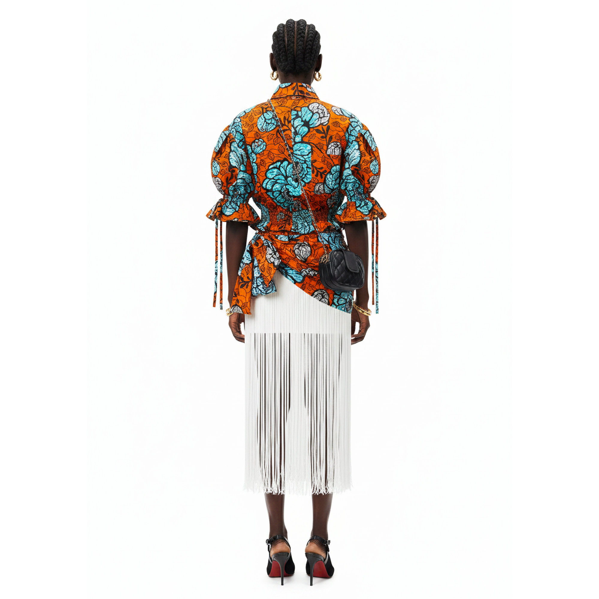 A model shown from the back wearing a vibrant orange and turquoise patterned wrap-style blouse with long fringe details, paired with a white fringed midi skirt and black high heels, against a solid white background.