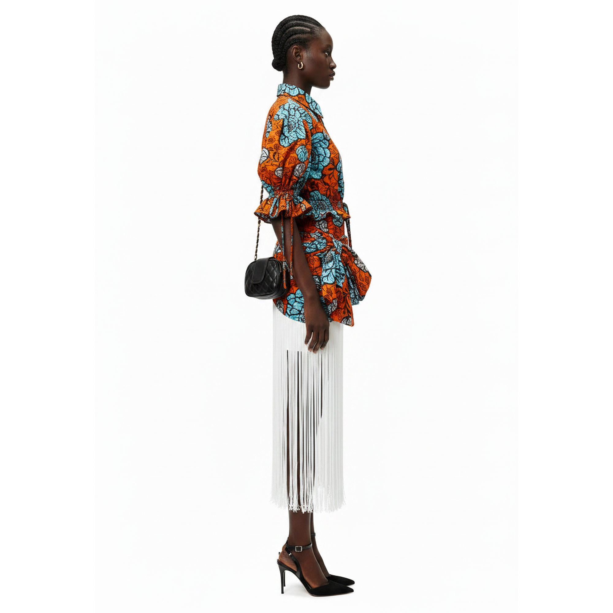 A model shown in profile wearing a vibrant orange and teal patterned shirt with a tie waist, paired with a white fringe midi skirt and black high heels, against a plain white background.