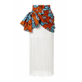 A white maxi skirt featuring a dramatic draped ruffle at the waist in a vibrant orange, blue, and black patterned African wax print, with a long white fringe cascading down the front.