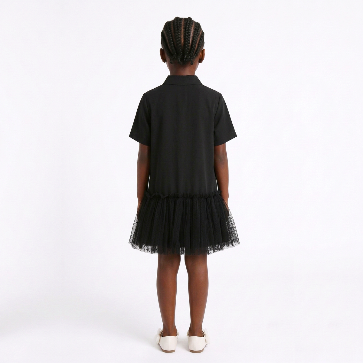 A child model shown from the back wearing a black short-sleeve collared shirt dress with a layered tulle skirt, paired with white slip-on shoes, against a plain white studio background.