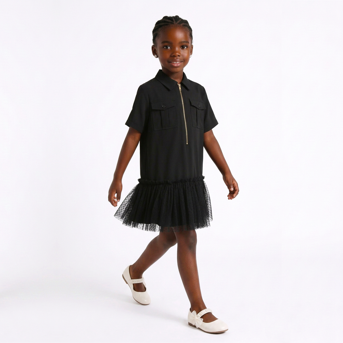 A young girl modeling a black short-sleeve collared dress with a gold zipper at the collar and a black tulle tutu skirt, paired with white Mary Jane shoes, against a plain white studio background.