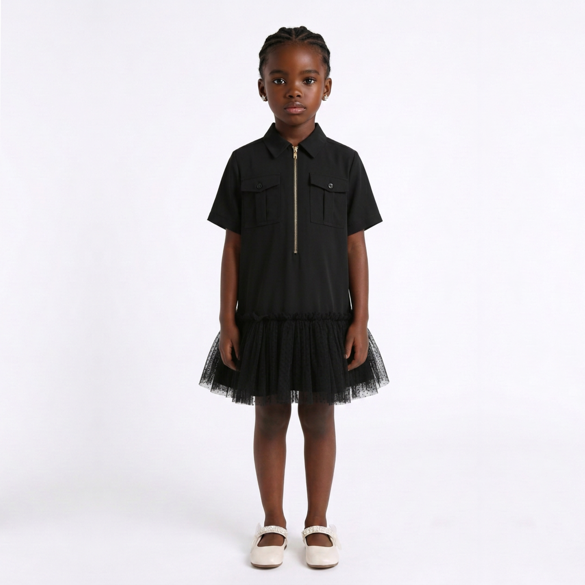 A young girl stands against a plain white studio background wearing a black short-sleeve dress with a front zipper, flap chest pockets, and a black tulle skirt. She is also wearing white Mary Jane shoes.