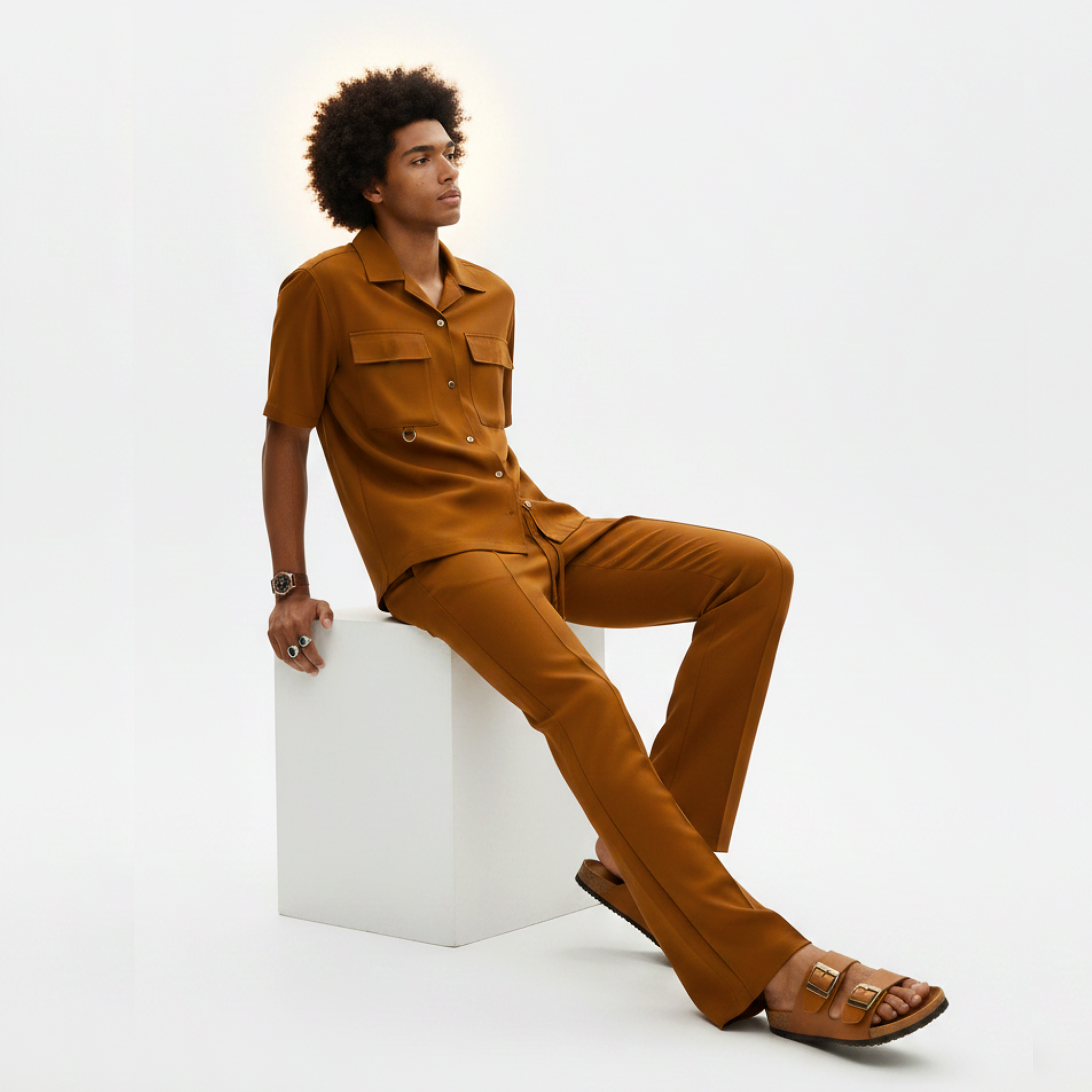 Man wearing a brown outfit sitting on a white cube against a white background