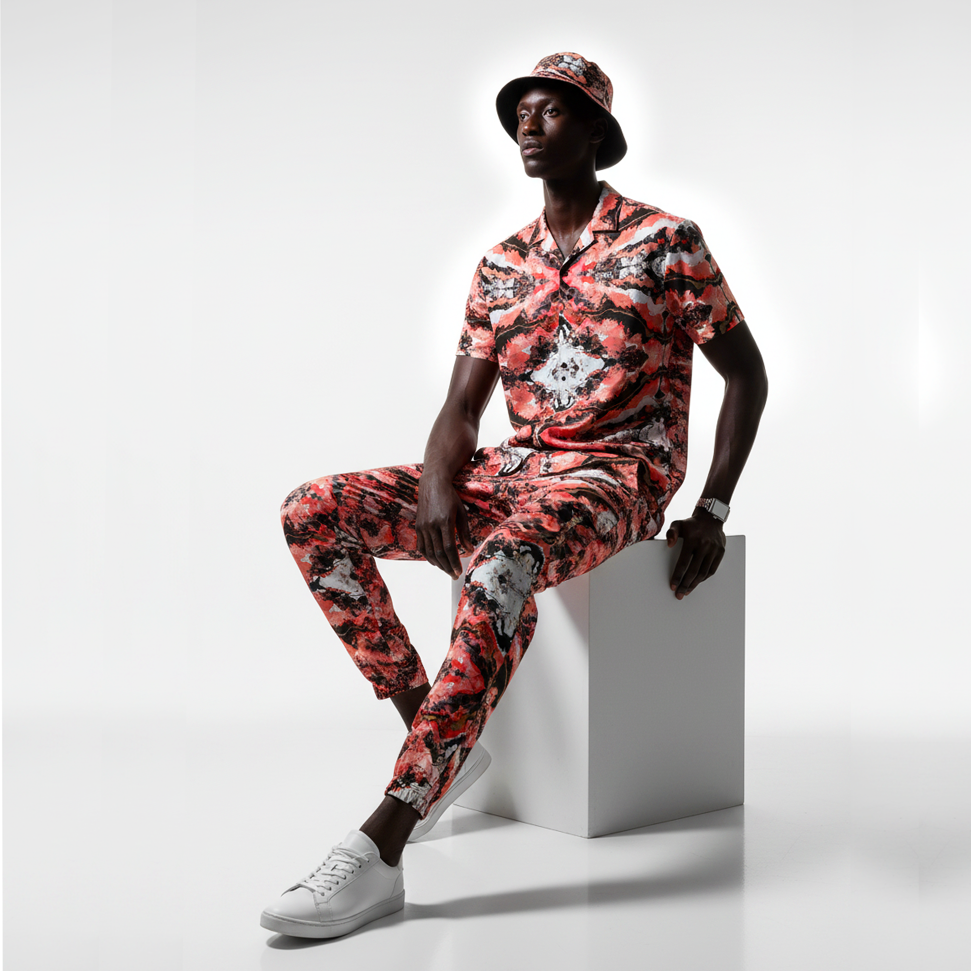 Man wearing a colorful floral outfit sitting on a white block against a light gray background