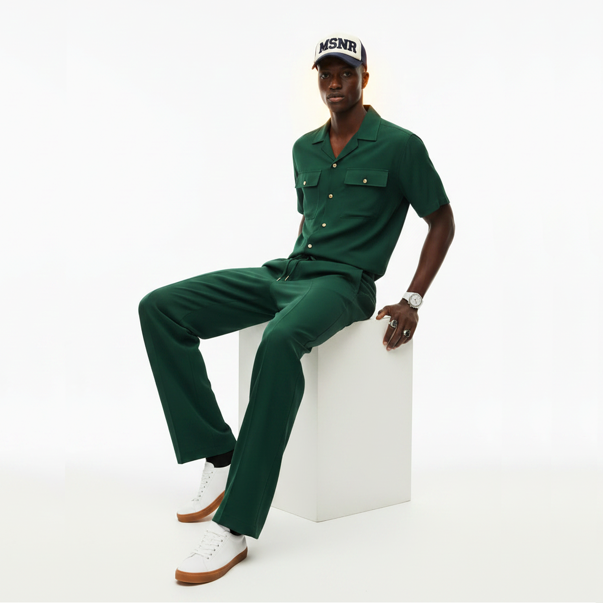 Man wearing a green outfit with a cap, sitting on a white cube against a white background