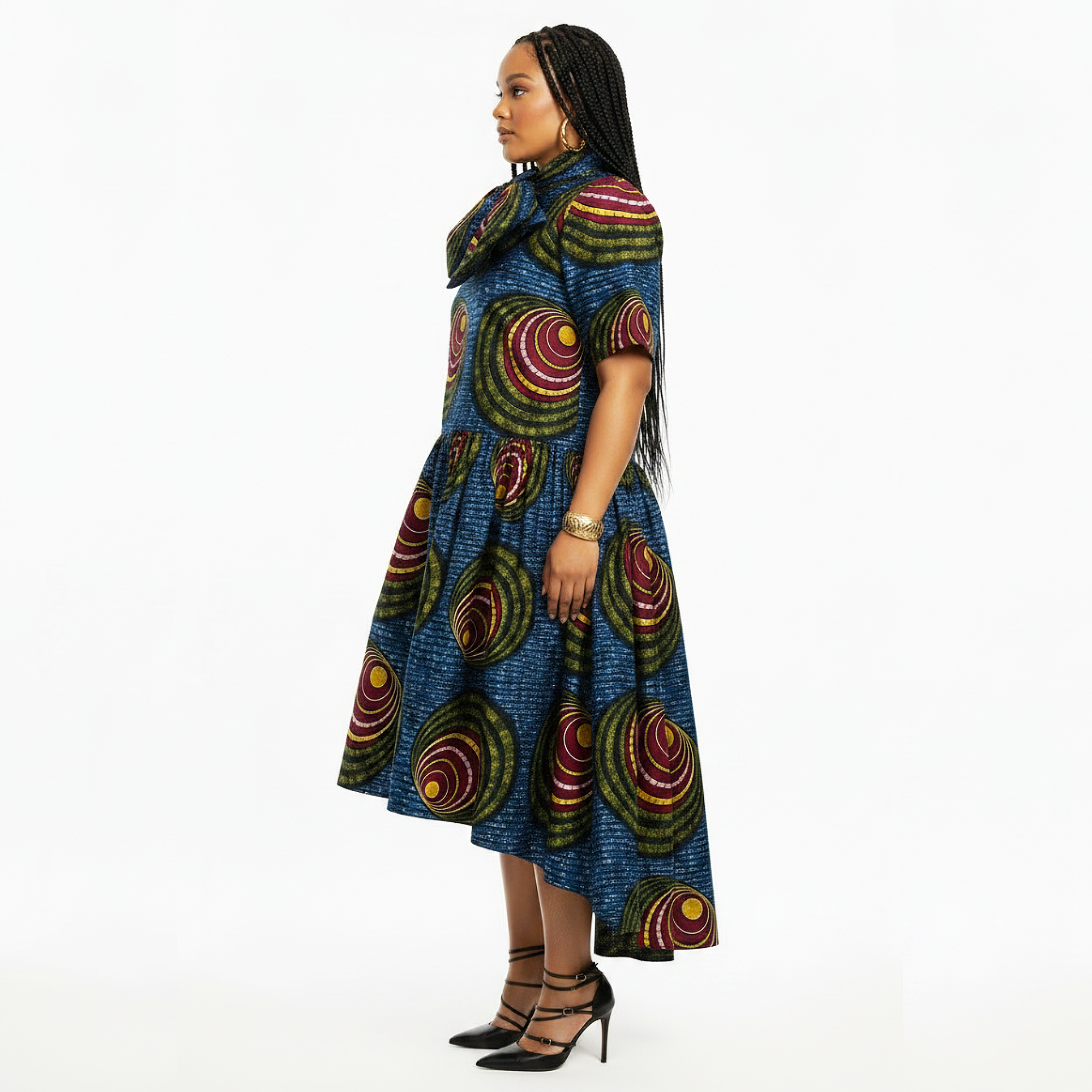 A woman modeling a short-sleeve high-low dress with a vibrant blue base and circular multicolor patterns, set against a plain white background.