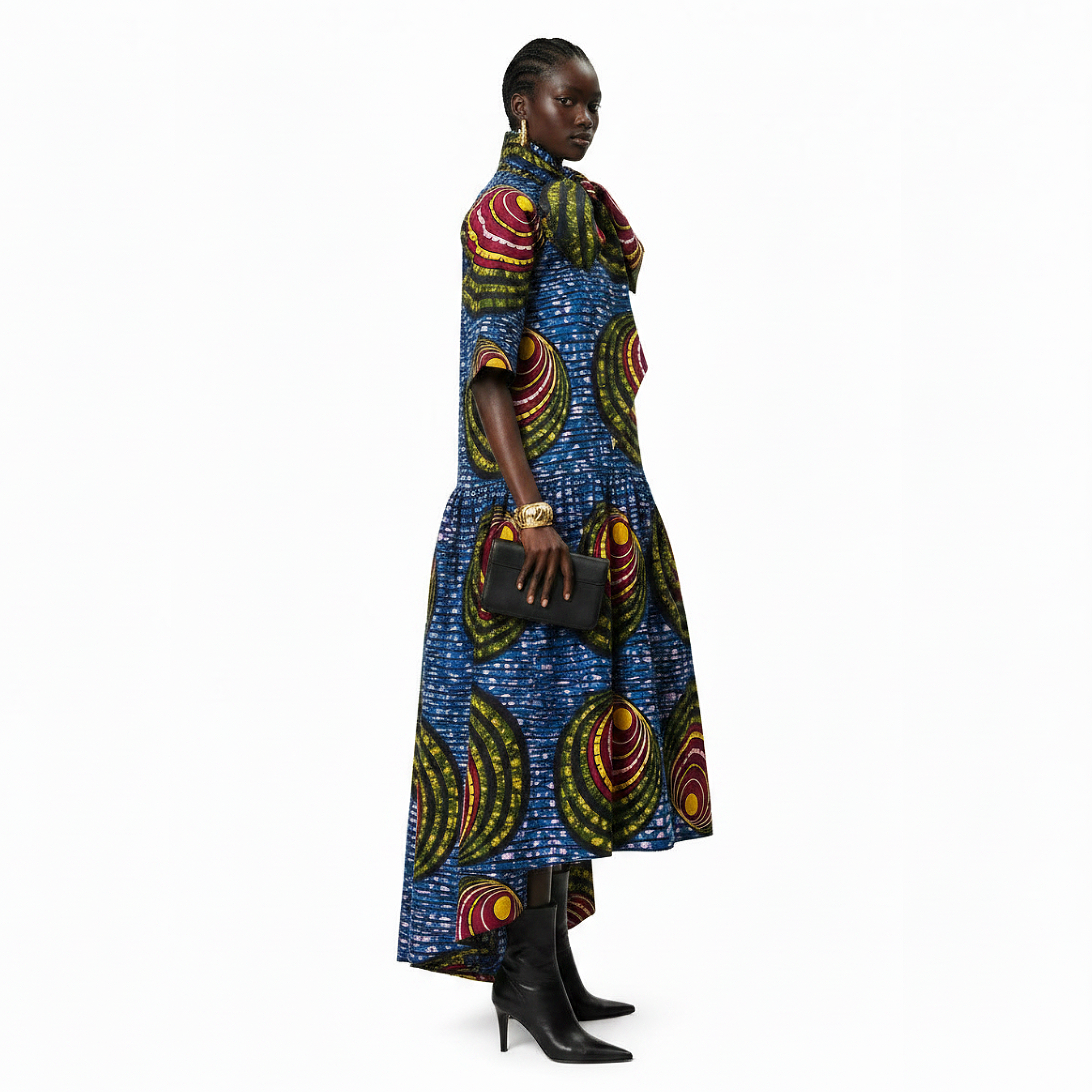 A model shown from the side profile wearing a vibrant, multicolored African wax print midi dress with a high collar, paired with black leather ankle boots and holding a black clutch, set against a plain white background.