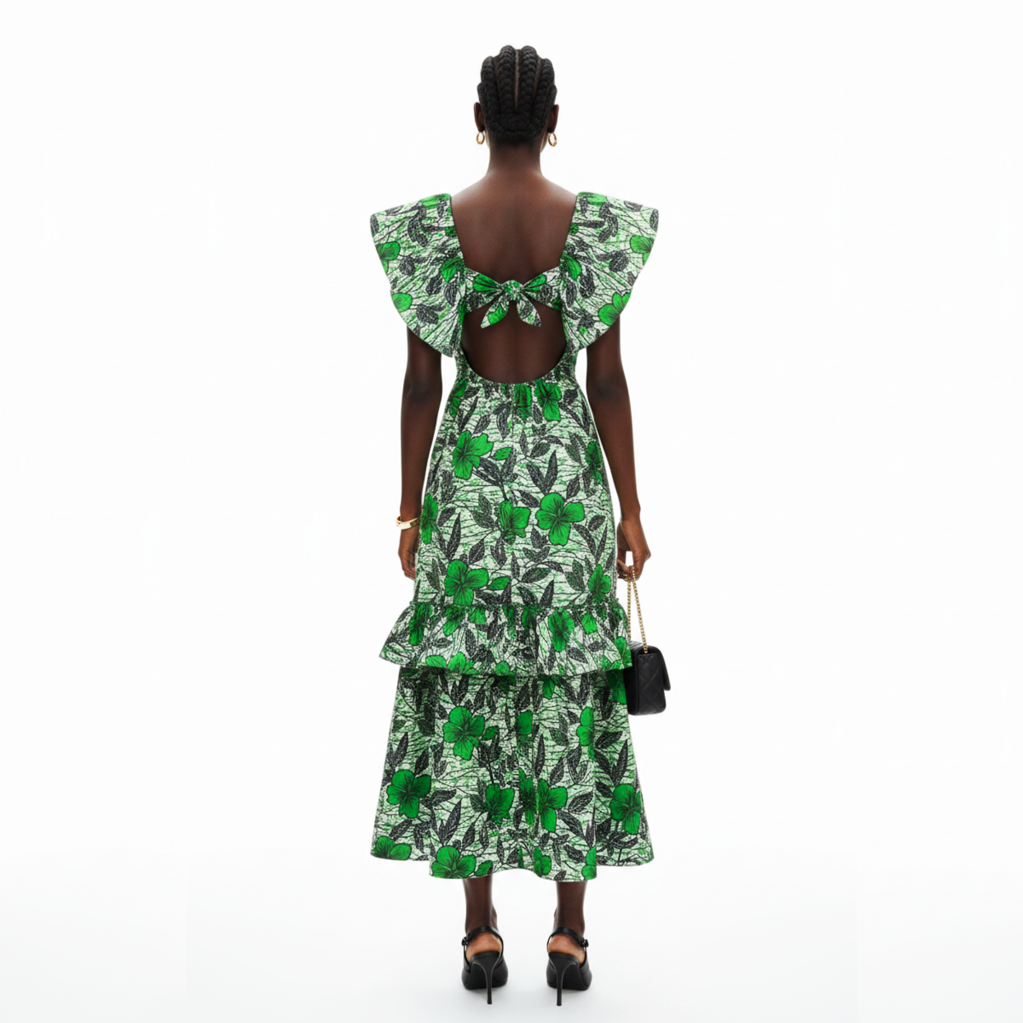 A woman viewed from the back wearing a green and white tropical floral print dress with ruffled shoulder details and an open back, paired with black heels and holding a small black handbag against a plain white background.