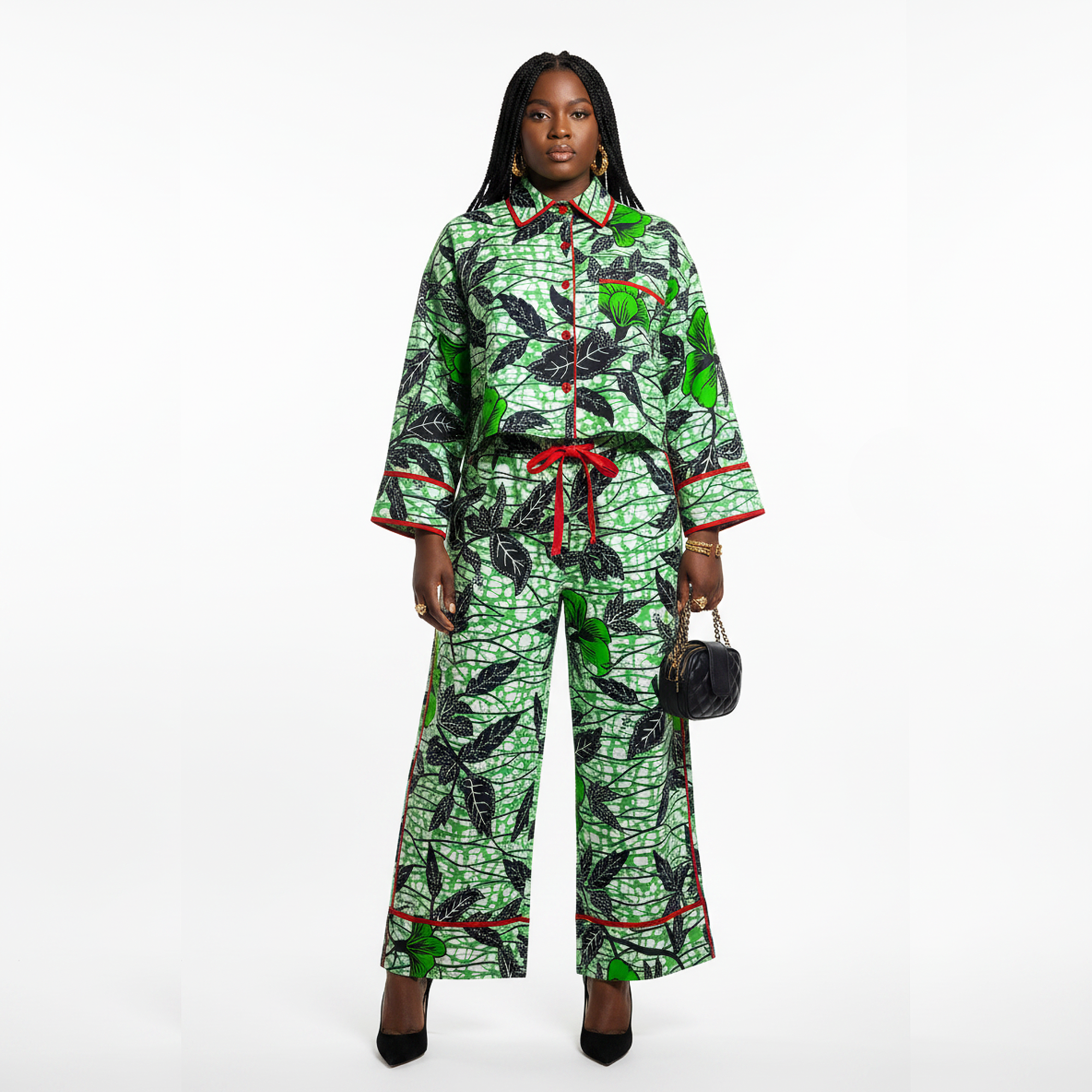 A model stands against a white studio background wearing a matching two-piece outfit consisting of a long-sleeve button-front top and wide-leg pants in a green and black leaf print with red trim and a red tie belt. She is holding a small black handbag.