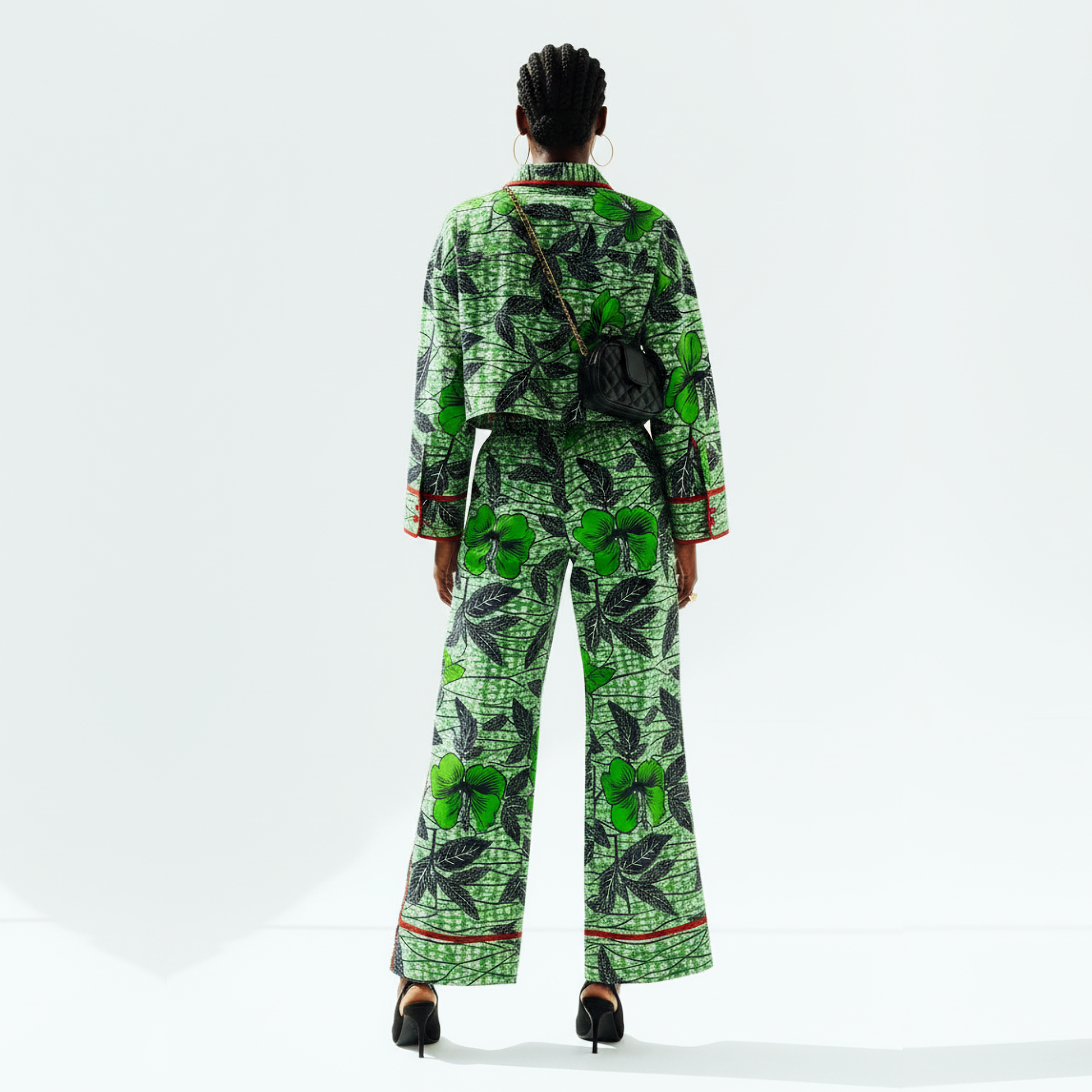 A model viewed from behind wearing a matching green and black patterned two-piece set with a long-sleeve top and wide-leg pants, styled with a small black crossbody bag and black heels against a white studio background.