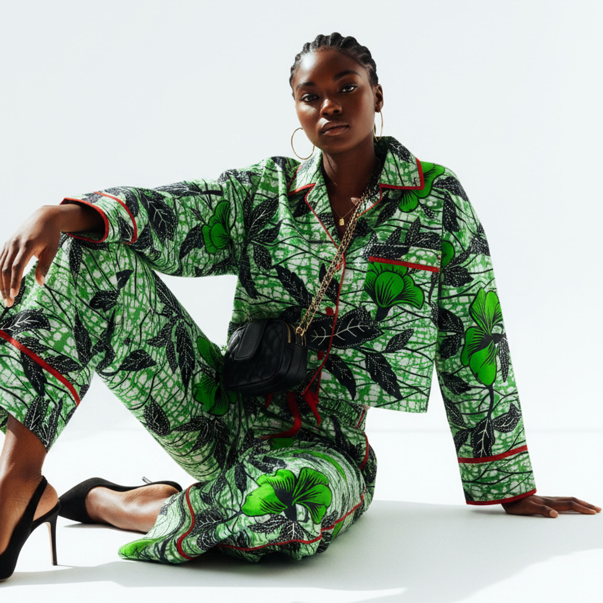 A model wearing a matching green and black patterned pajama set with red piping, sitting on a white studio floor.