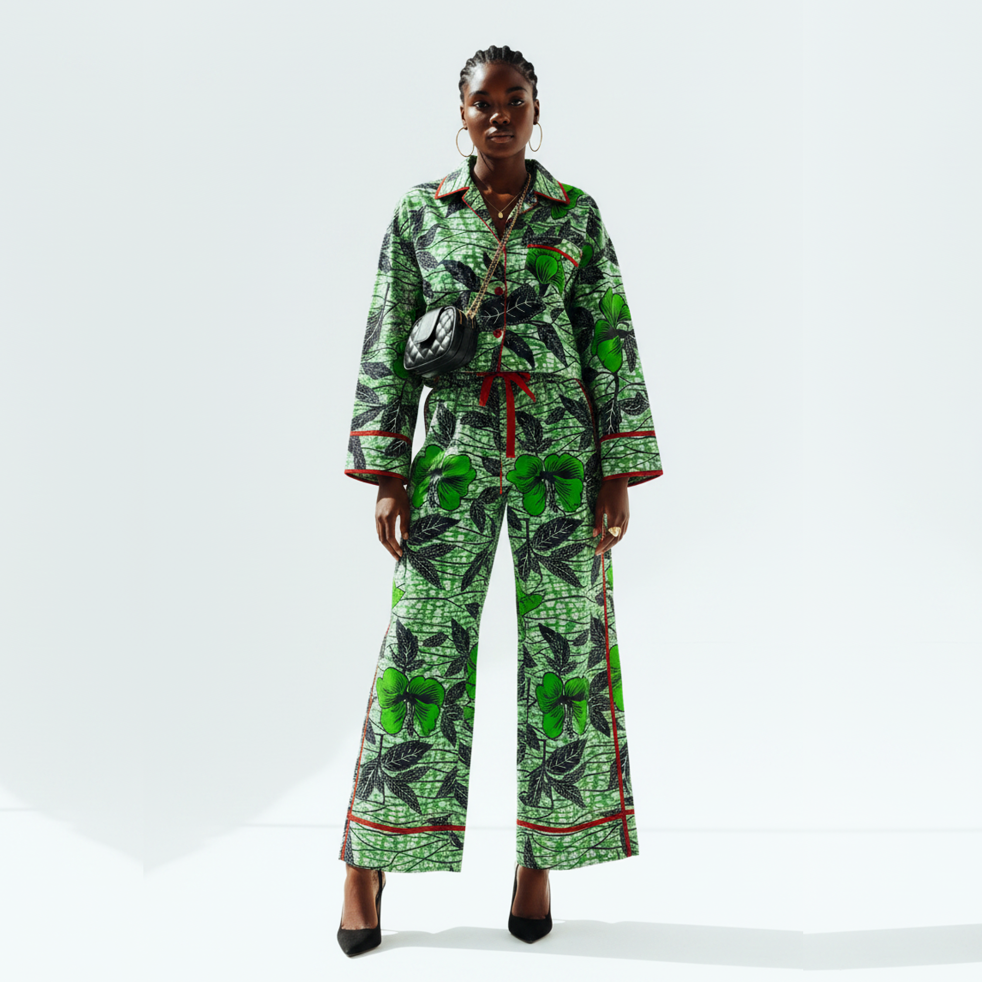 A model stands against a light gray studio background wearing a matching two-piece pajama set with a green and black leaf pattern and red trim details.