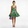 A model stands against a white studio background wearing a sleeveless fit-and-flare dress with a bold green, black, and red abstract print, holding a purple handbag.