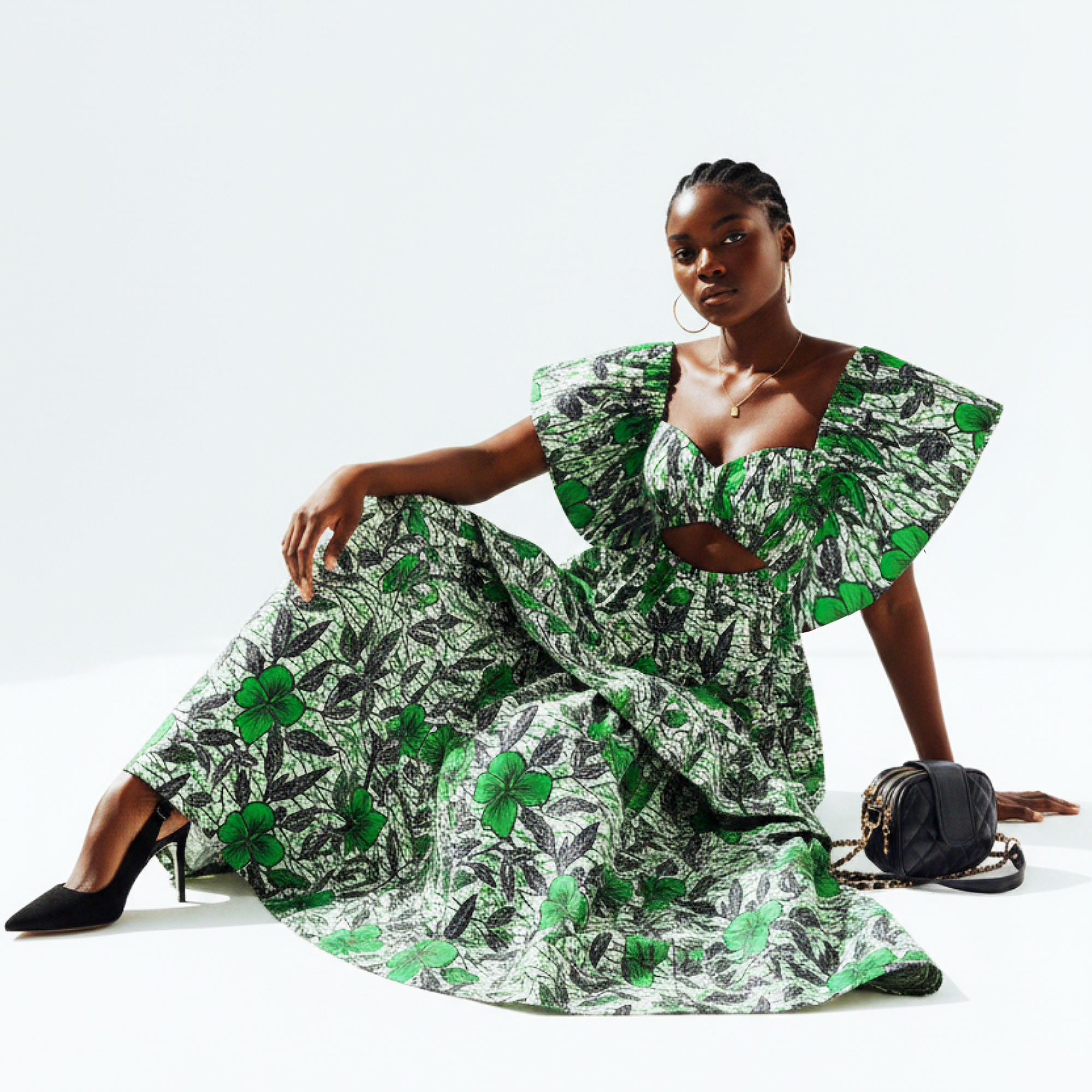 A model wearing a two-piece matching set consisting of a ruffled crop top and wide-leg trousers, both in a green and black tropical floral print on a white background. She is seated on a white studio floor, wearing black high heels and holding a small black handbag.