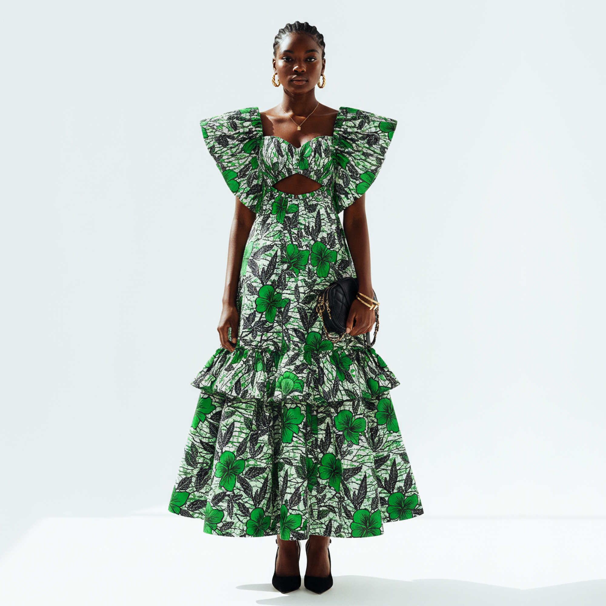 A model stands against a light gray studio background wearing a long tiered dress with a bold green and black botanical print, ruffled cap sleeves, a square neckline with a front cutout, and black pointed heels.
