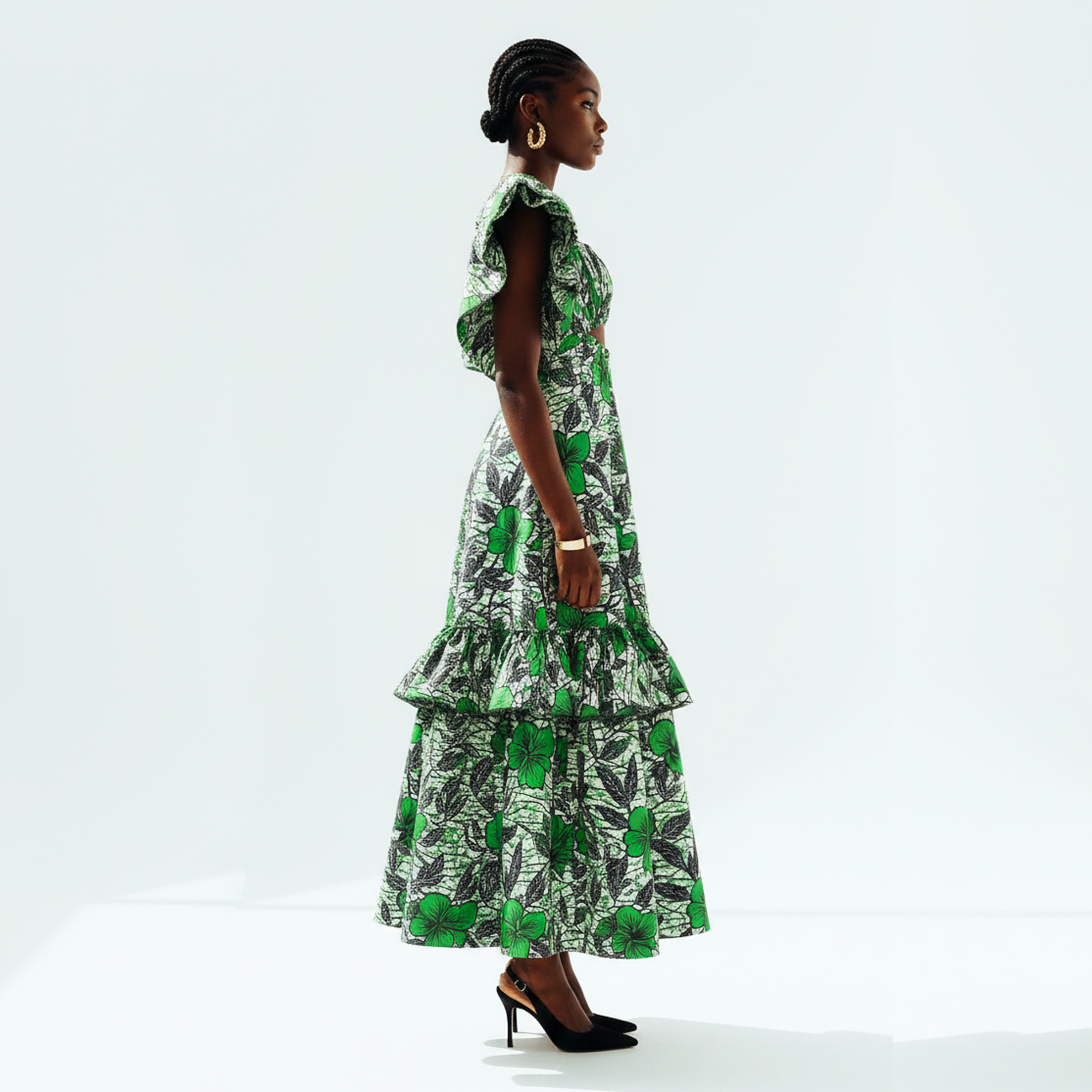 A model is shown from the side profile wearing a sleeveless tiered maxi dress with a green and black botanical leaf print, styled with black heels against a clean white studio background.