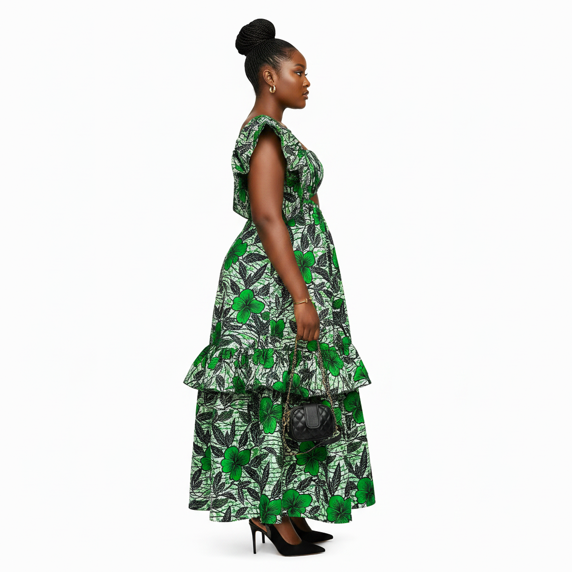 A woman stands in profile wearing a sleeveless, tiered maxi dress with a green and black tropical leaf print. She is holding a small black handbag and wearing black high heels, photographed against a solid white studio background.
