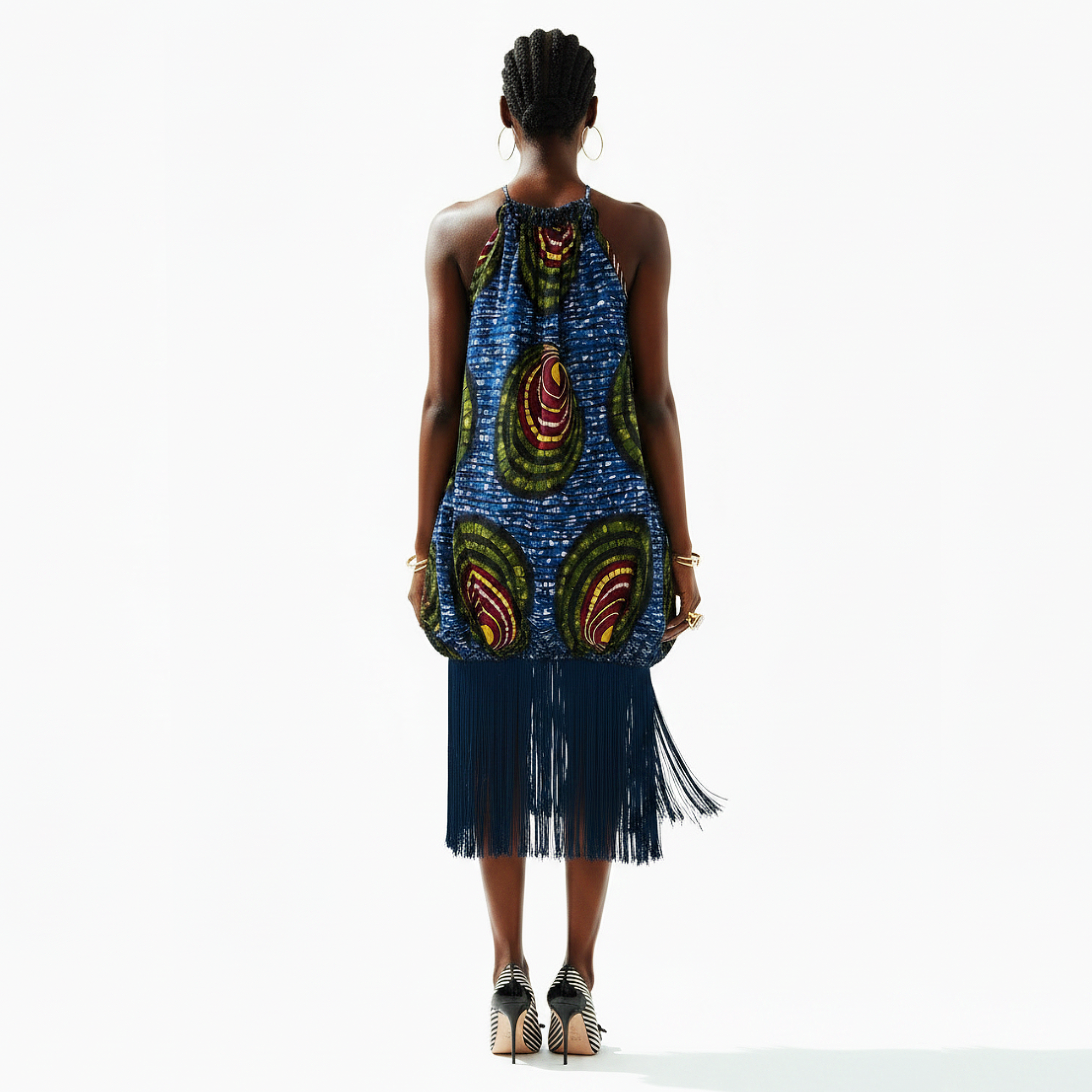 A model is shown from the back wearing a sleeveless halter dress with a blue and green African wax print pattern and a long dark fringe hem, paired with black and beige high-heeled sandals against a white studio background.