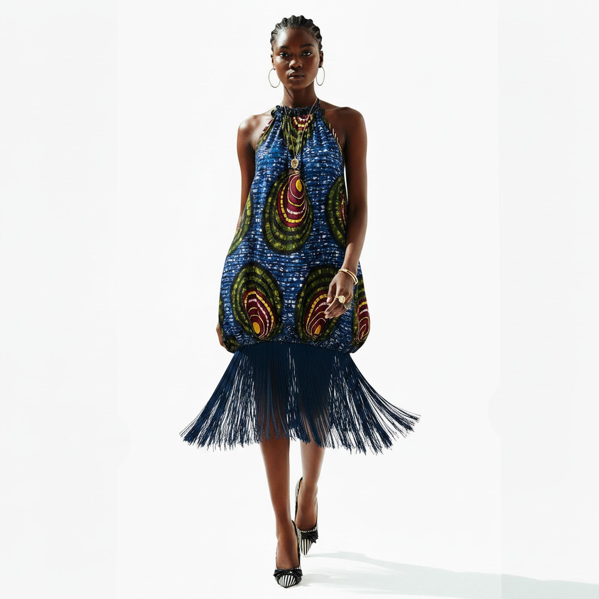 A model walks against a white studio background wearing a sleeveless halter-neck dress with a vibrant blue, green, and red abstract pattern and a dark navy fringe hem.