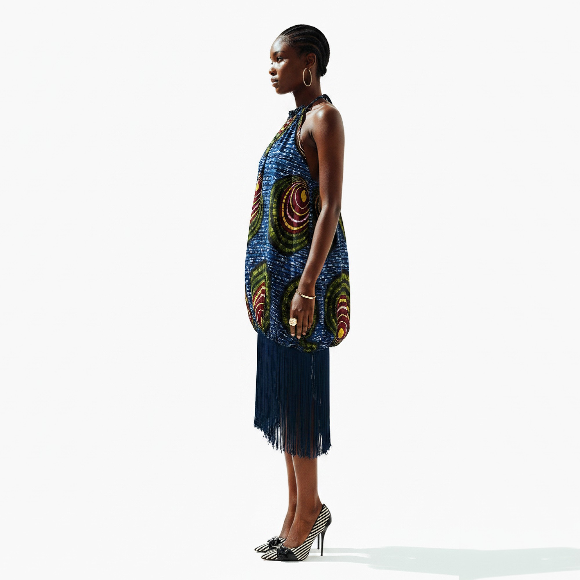 A model is shown in profile wearing a sleeveless halter dress with a blue base and vibrant multicolor circular patterns, featuring a long dark blue fringe skirt, set against a plain white studio background.