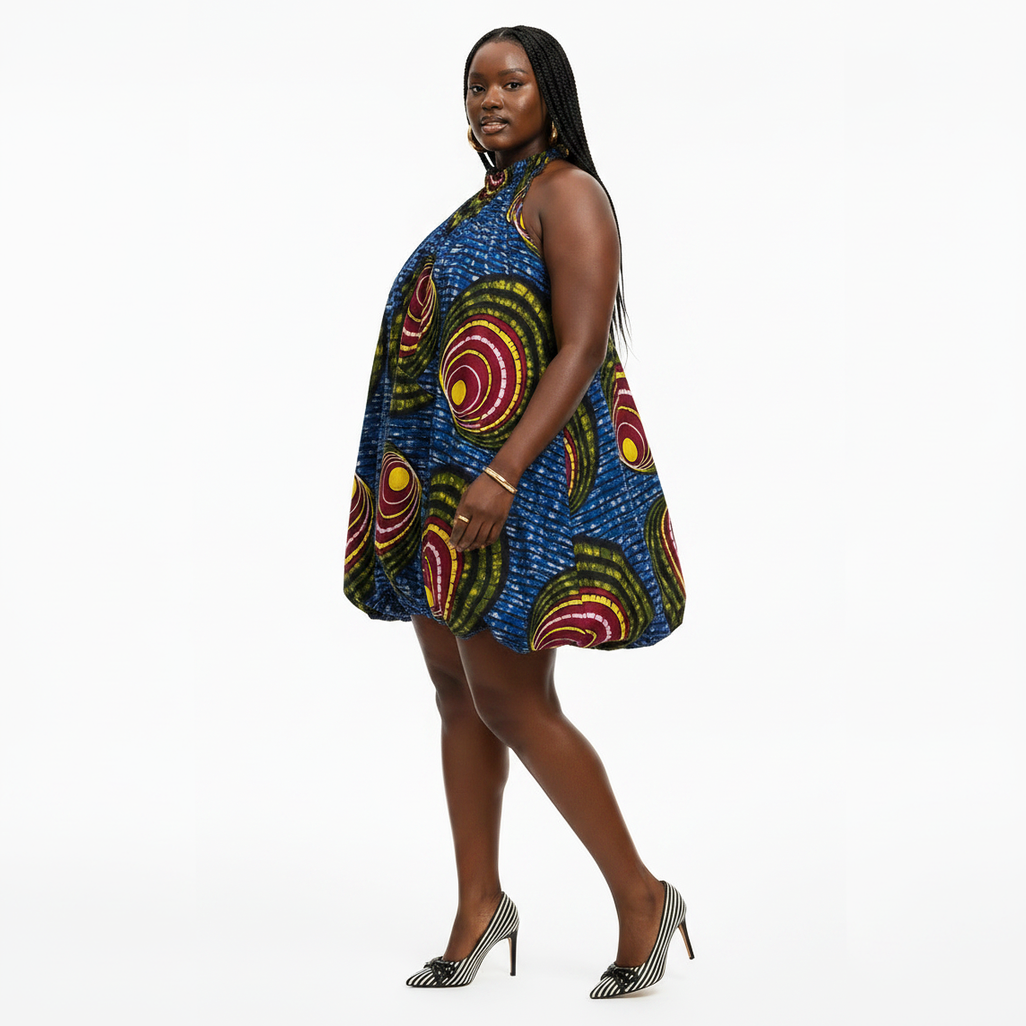 A woman modeling a sleeveless halter-neck mini dress with a vibrant blue base and a bold multicolor circular pattern in red, yellow, and green, paired with black and white striped high heels against a plain white studio background.