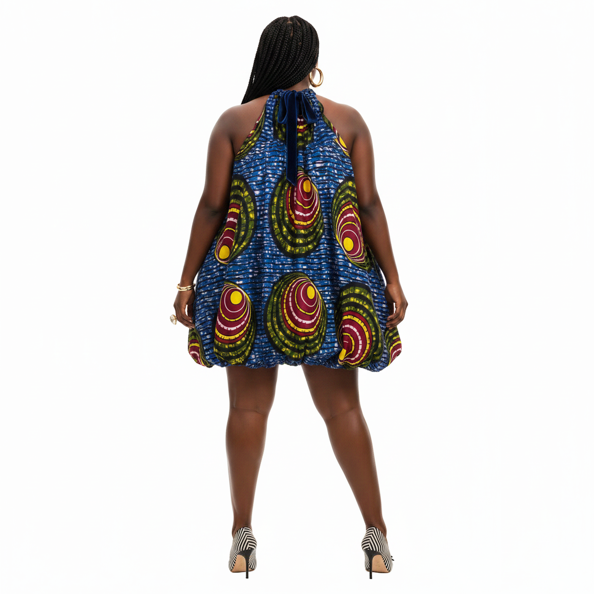 A woman viewed from the back wearing a sleeveless halter mini dress with a vibrant blue base and large circular multicolor patterns in red, yellow, and green. She is standing against a solid white background.