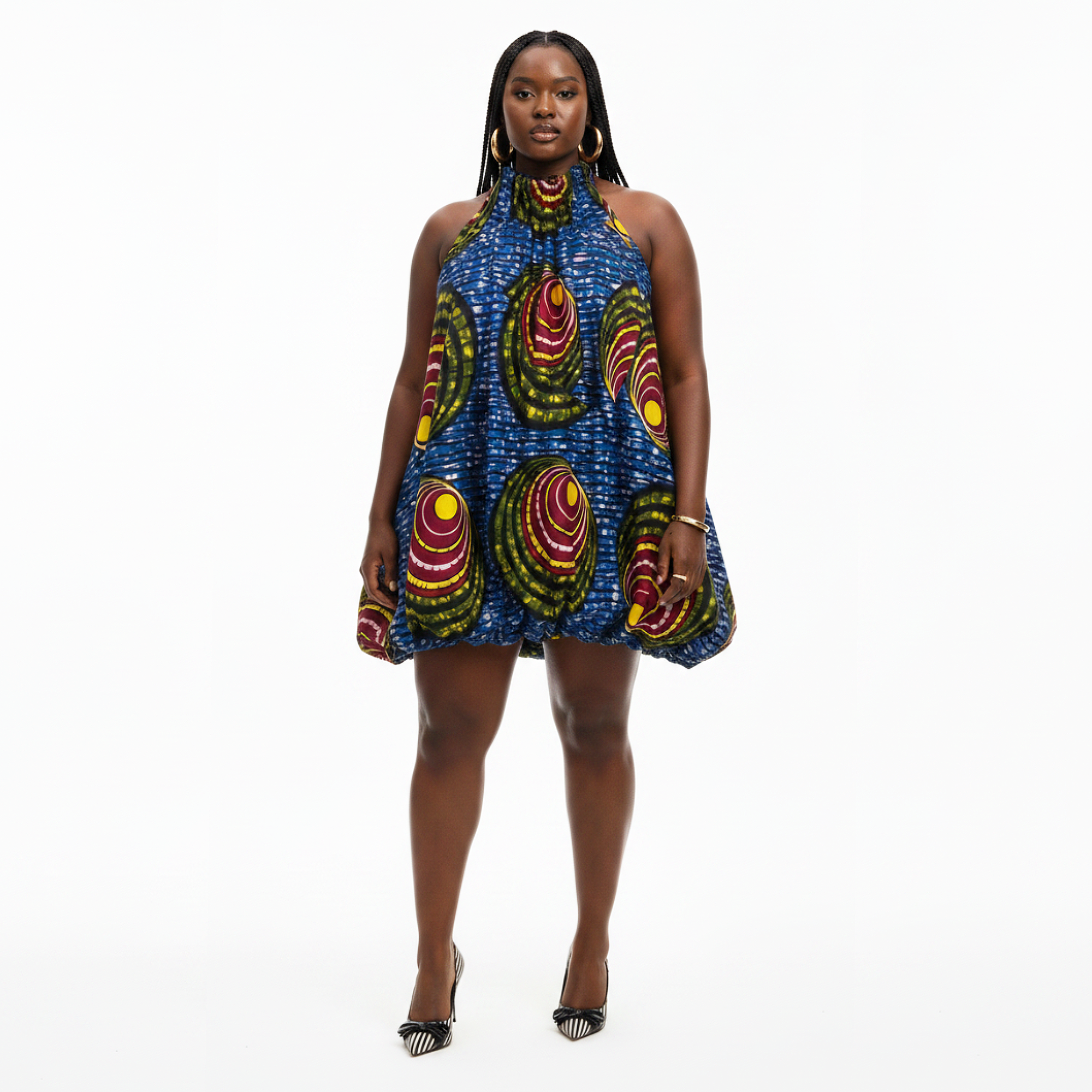 A woman modeling a sleeveless halter-neck dress with a vibrant blue base and large circular African wax print motifs in red, yellow, and green, paired with black high heels against a white studio background.