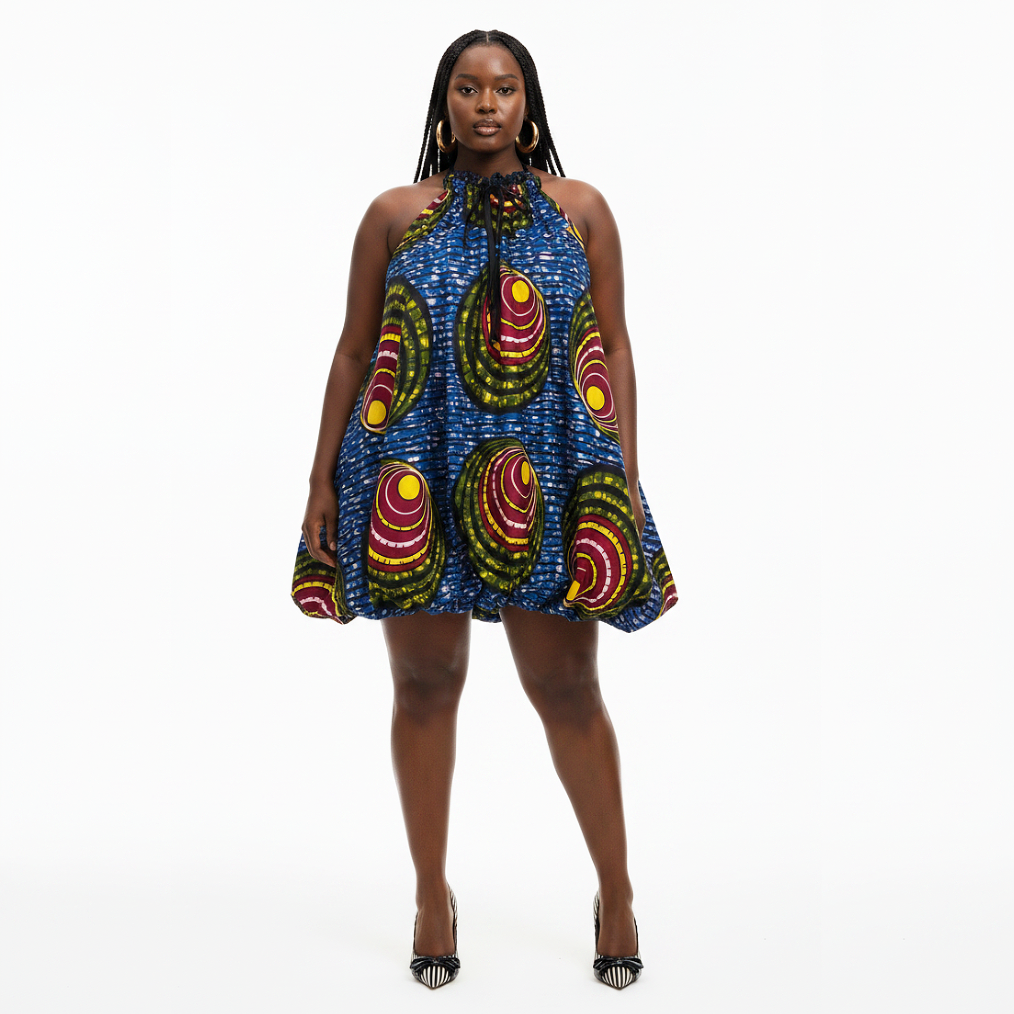 A woman modeling a sleeveless halter-neck mini dress with a vibrant blue African wax print pattern featuring red, yellow, and green concentric circles, set against a plain white studio background.