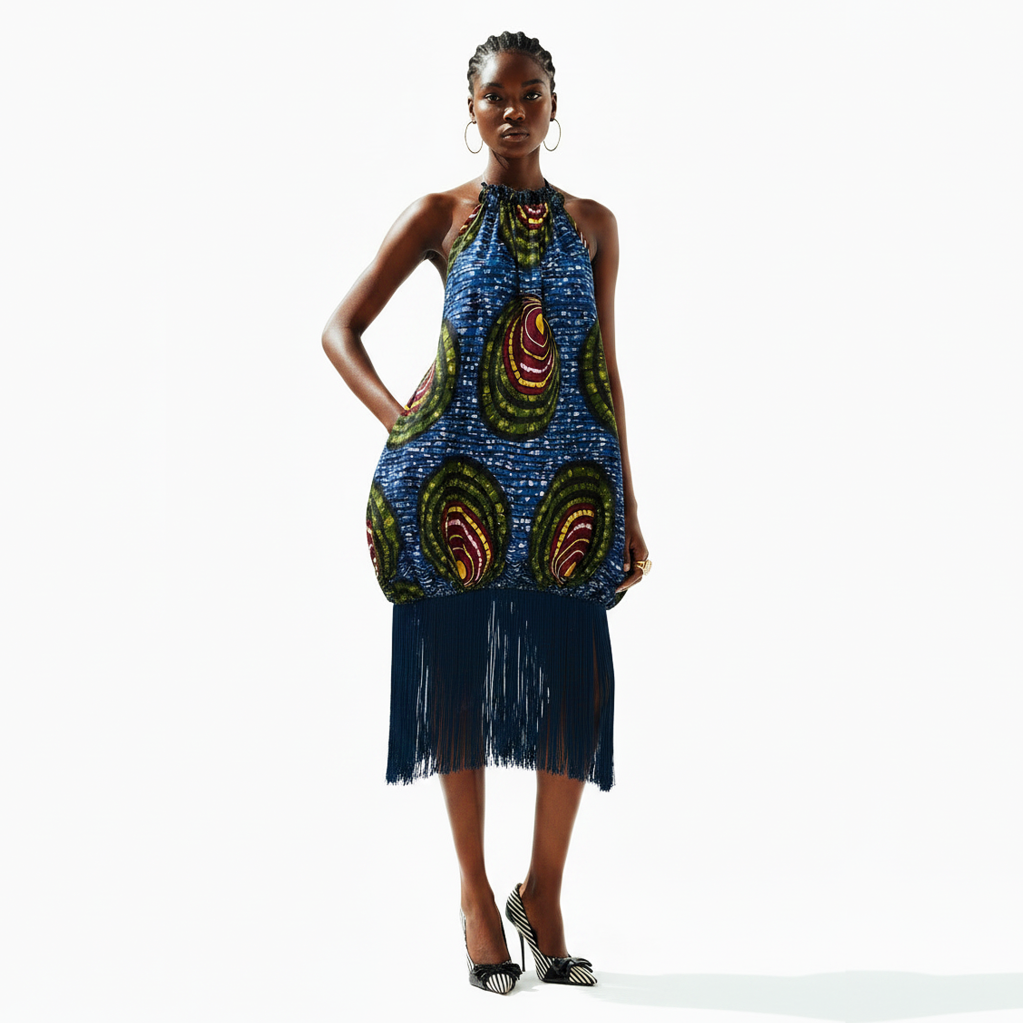A model stands against a plain white studio background wearing a sleeveless halter dress with a blue base and large multicolor paisley-style motifs in green, red, and yellow, plus a long dark fringe skirt and black high heels.