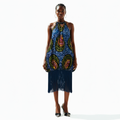 A model stands against a white studio background wearing a sleeveless halter-neck dress with a blue base and large multicolor paisley-like motifs, finished with a long dark fringe hem. She is also wearing hoop earrings and black high heels.
