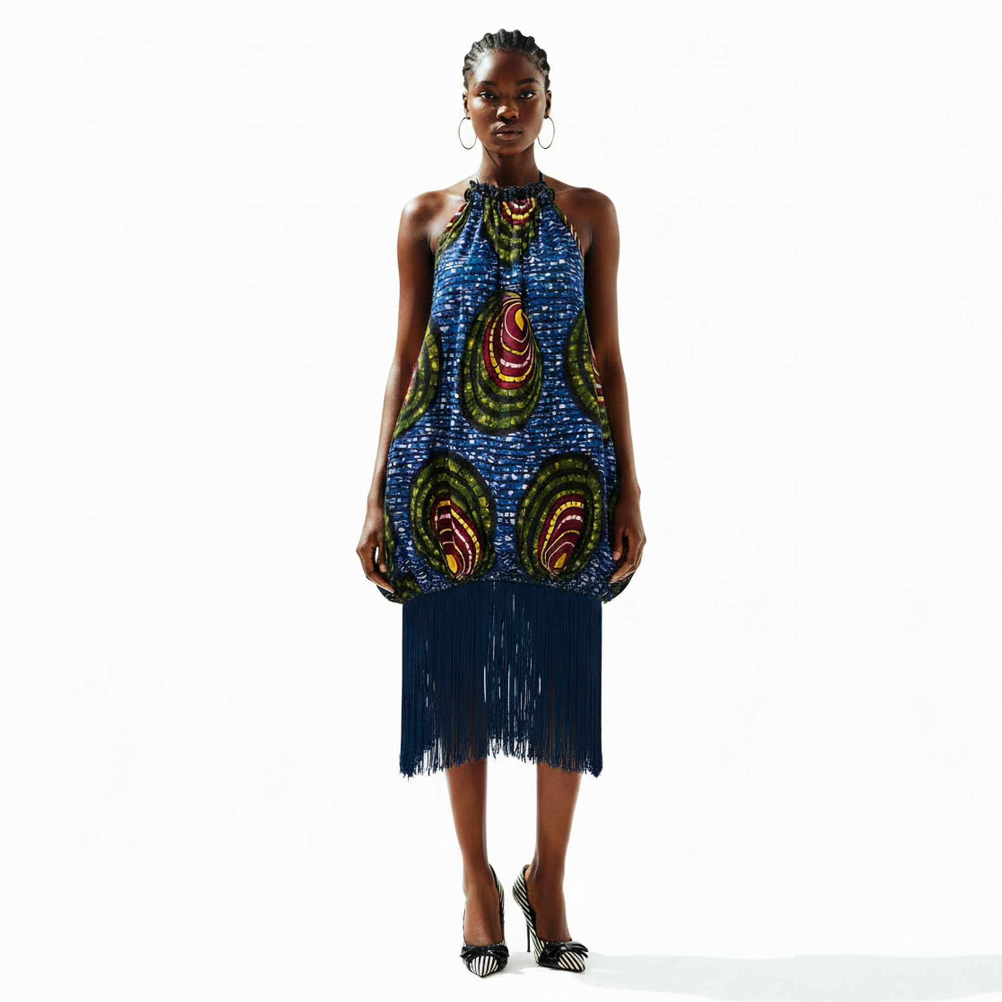 A model stands against a white studio background wearing a sleeveless halter-neck dress with a blue base and large multicolor paisley-like motifs, finished with a long dark fringe hem. She is also wearing hoop earrings and black high heels.