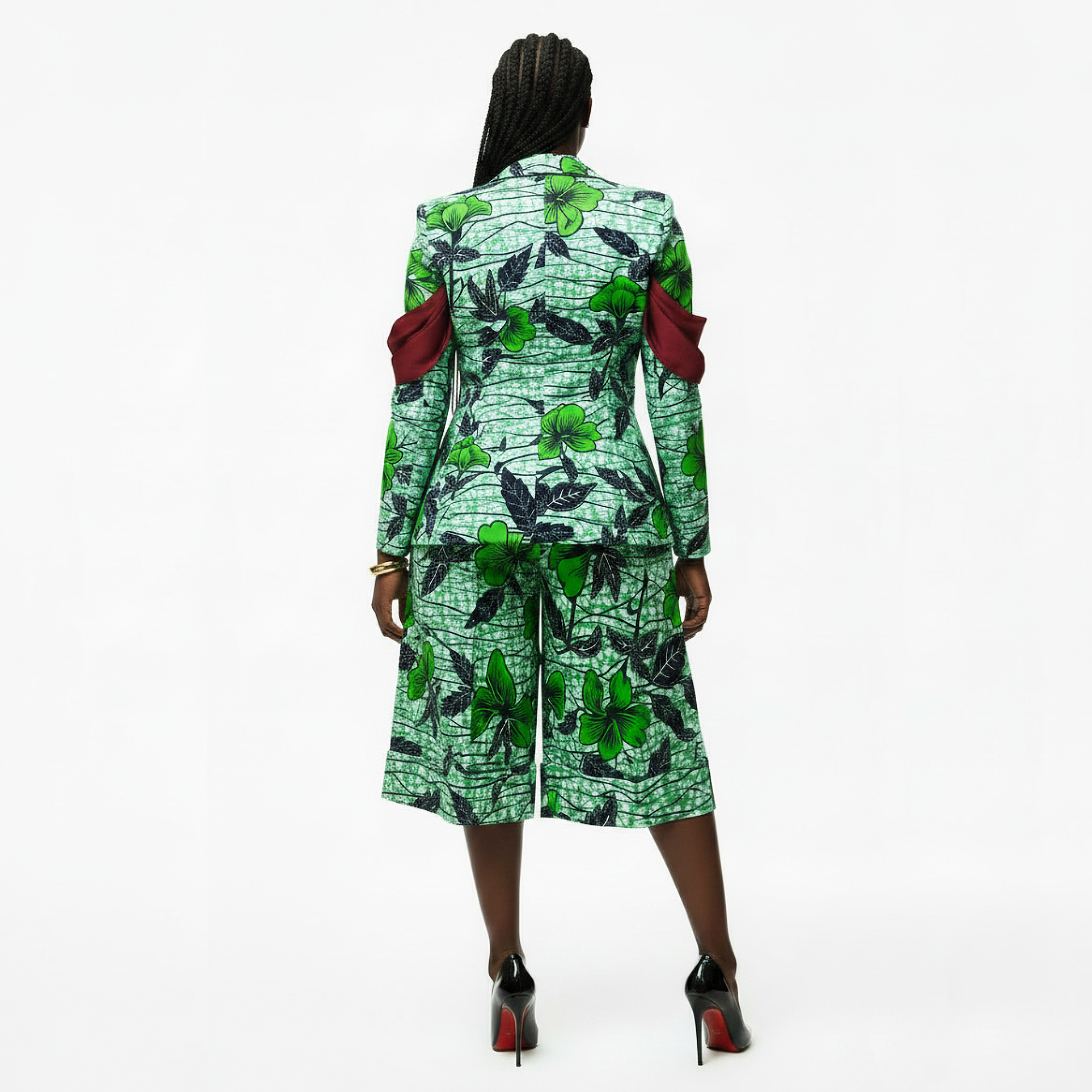 A model viewed from the back wearing a green and black floral print two-piece suit with contrasting dark red shoulder details, paired with black high heels, on a white studio background.