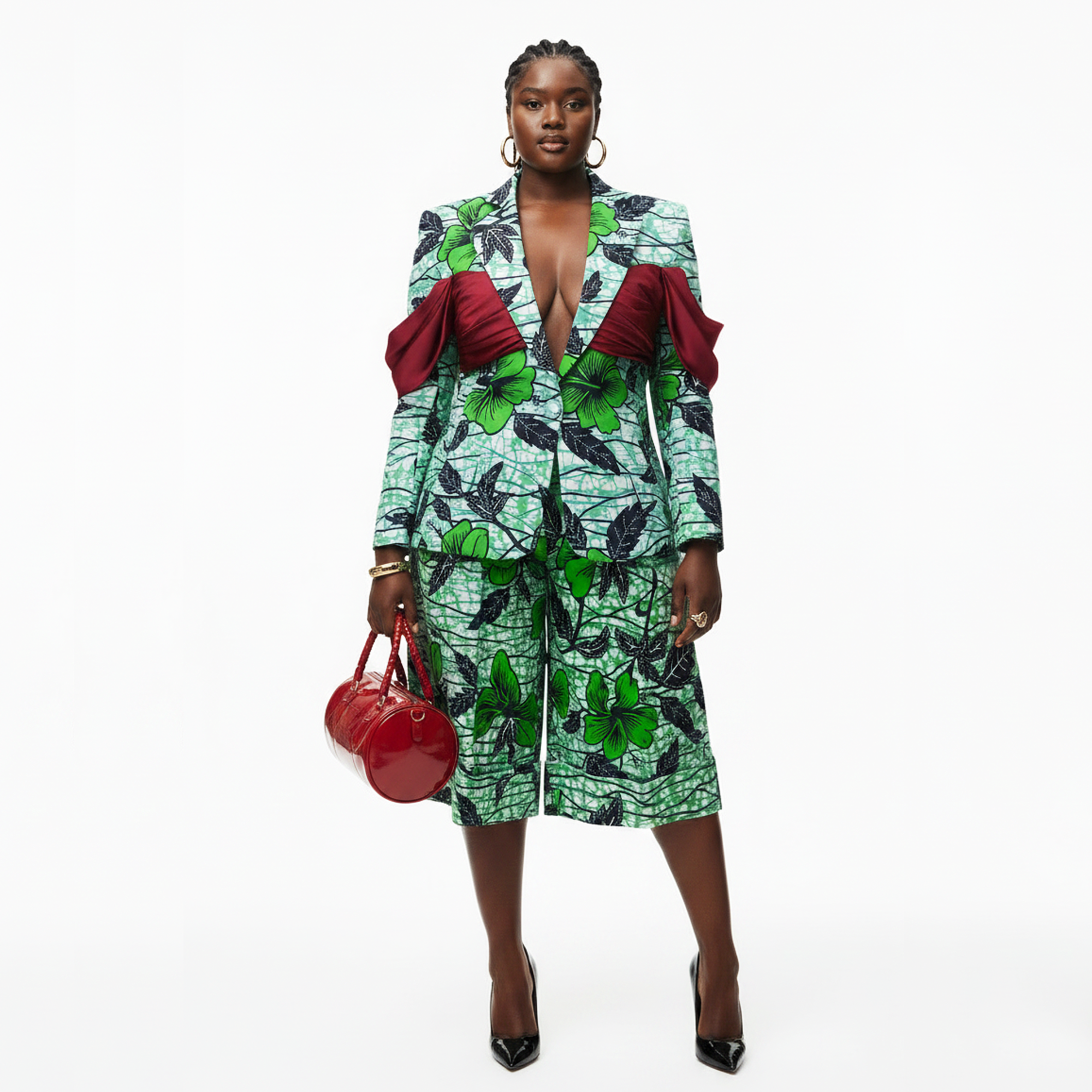 A model stands against a white studio background wearing a green and black leaf-patterned blazer and matching skirt with bold red draped shoulder details, holding a red handbag and wearing black pointed heels.