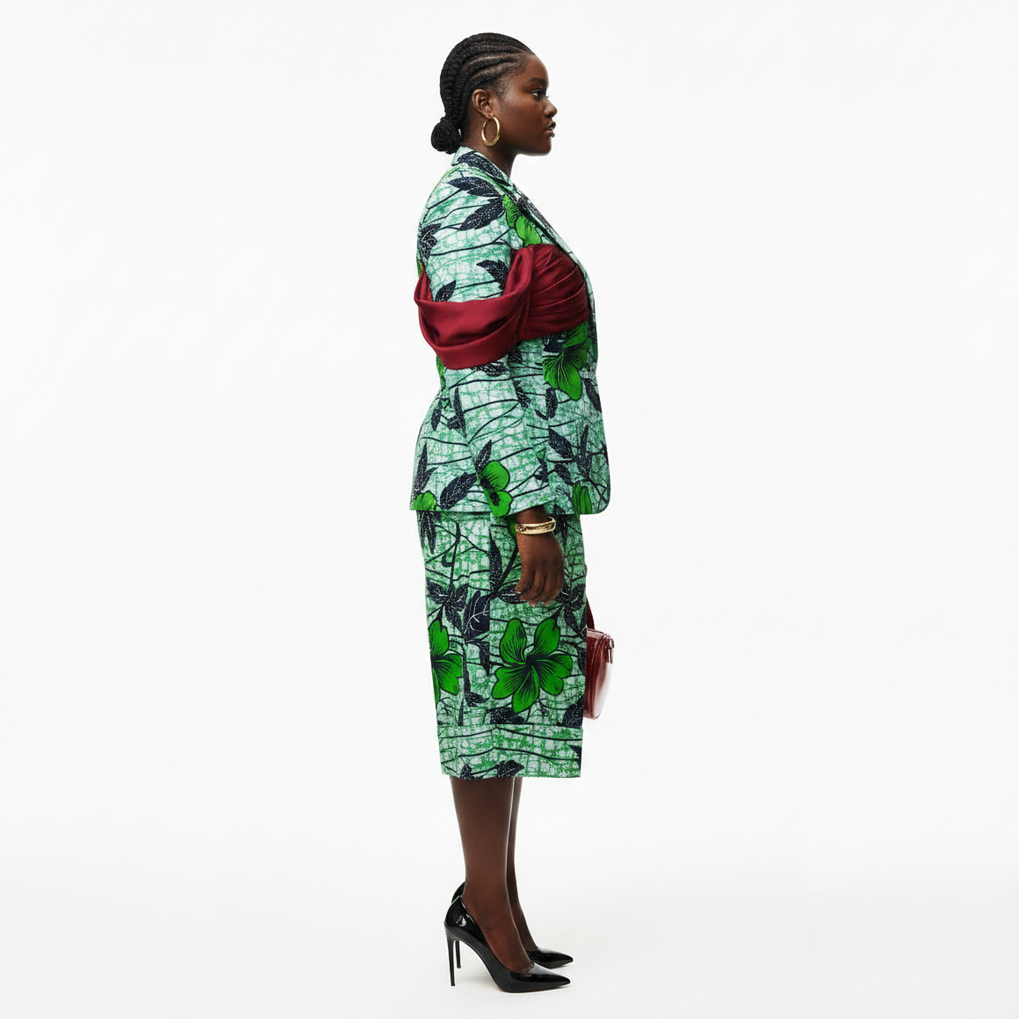 A woman stands in profile wearing a green and black patterned dress with a draped red sash at the waist, paired with black high heels, against a plain white studio background.