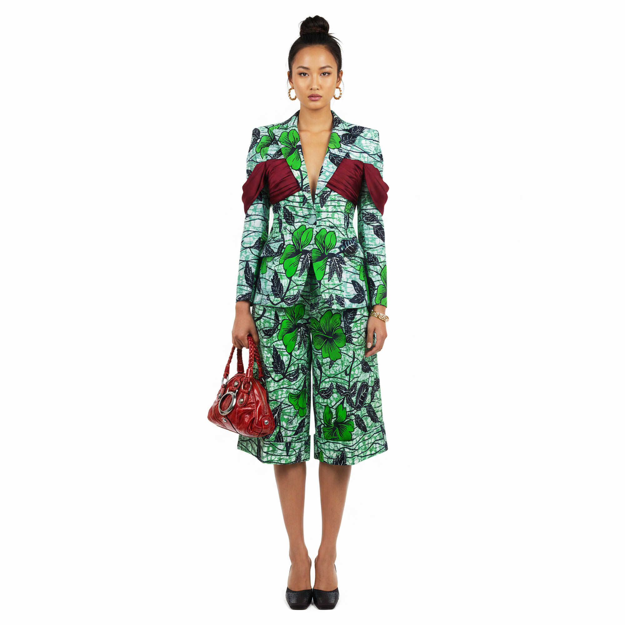 A model stands against a white background wearing a green and black patterned blazer and matching midi skirt with dark red ruffled shoulder details, holding a red handbag.