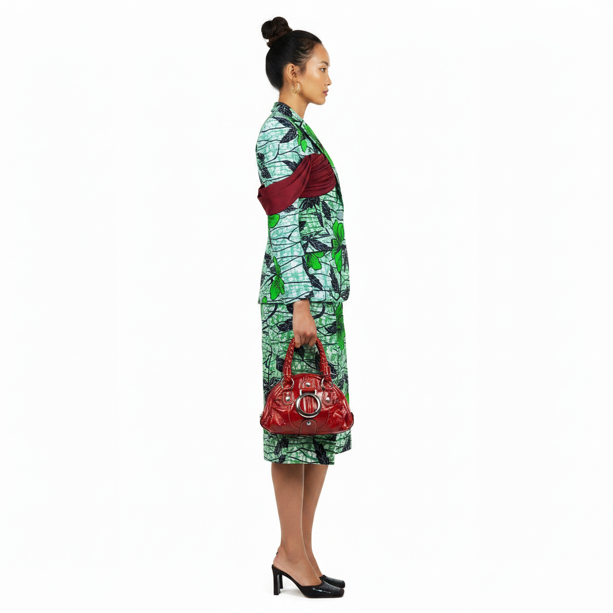 A woman stands in profile wearing a green and white patterned matching set with a long-sleeve top and skirt, holding a red handbag against a plain white background.