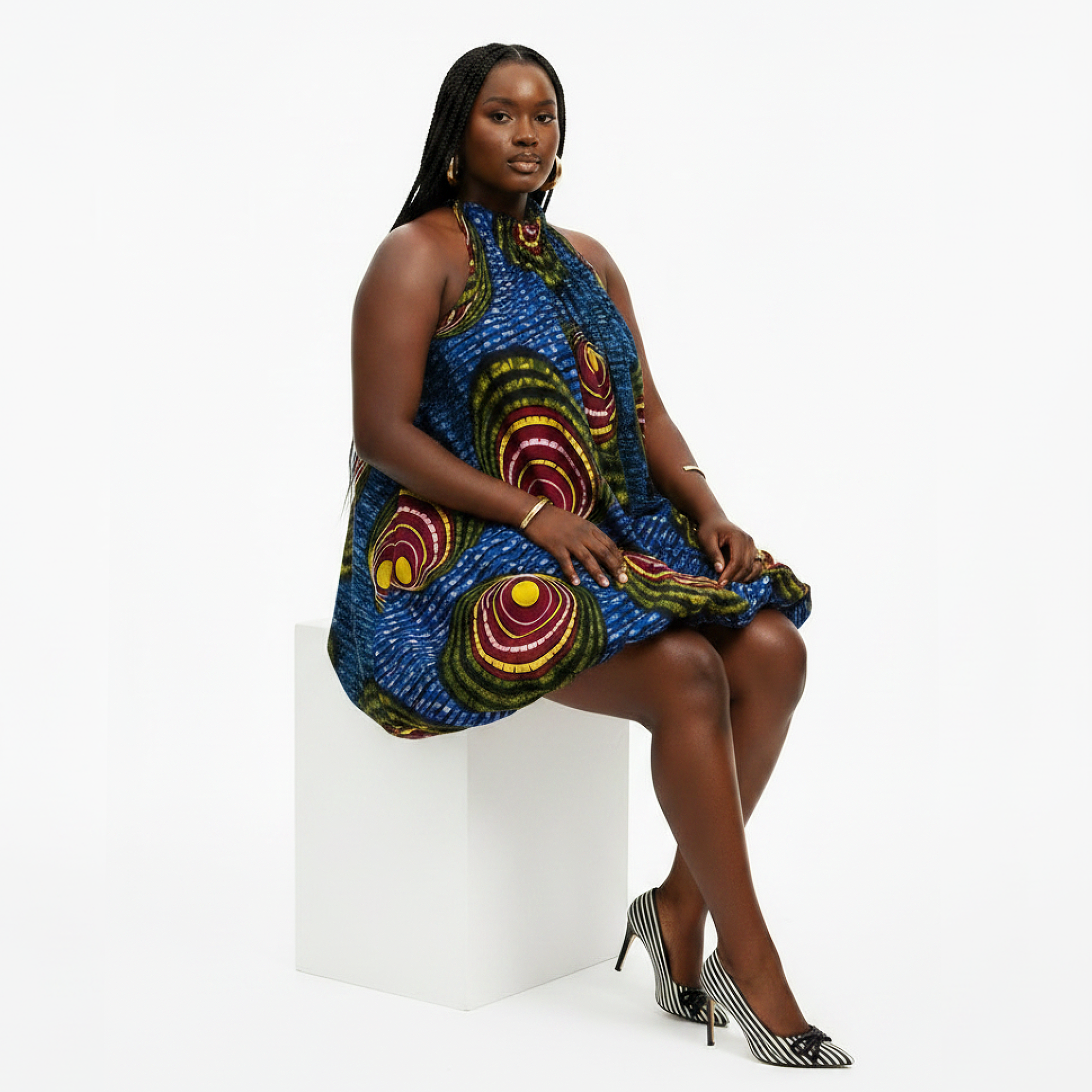 A woman models a sleeveless high-neck dress with a vibrant blue base and large circular African wax print motifs in red, yellow, and green, seated on a white block against a white studio background.